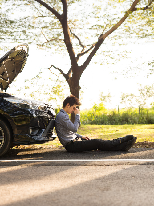 businessman with hands on head sitting by broken down car