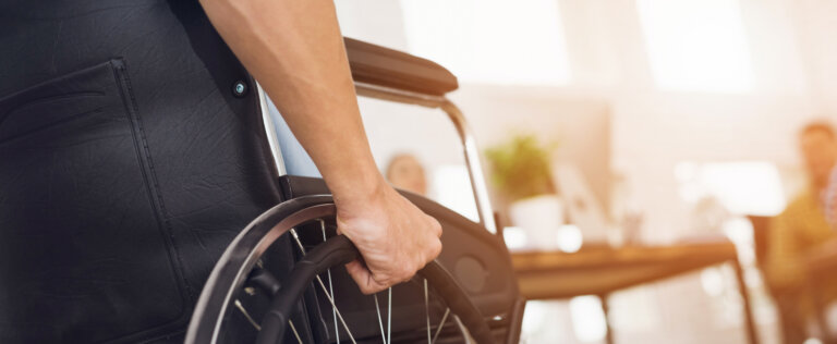 A disabled man is sitting in a wheelchair. He holds his hands on the wheel. Nearby are his colleagues
