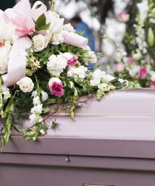 "Pink casket adorned with a large floral arrangement of white and pink roses, tulips, carnations, and greenery, with a pink ribbon on top during a funeral service."
