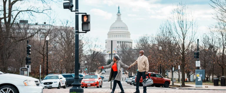 A vibrant senior couple of African American Descent hold hands and wait at a crosswalk while exploring Washington DC on a winter vacation, with a view of the United States Capitol visible in the background.