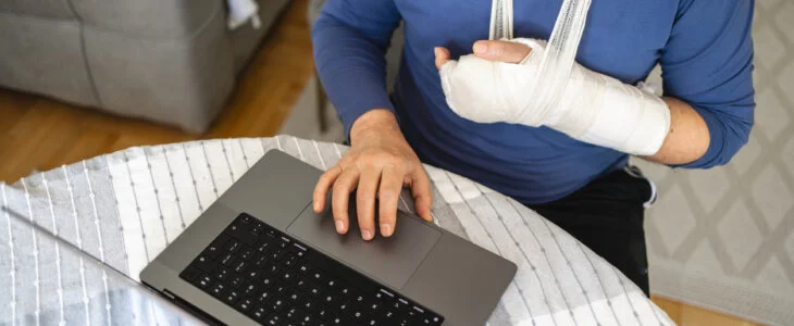 Unrecognizable senior or mid-adult Caucasian man, using a laptop while having a hand in the orthopedic cast